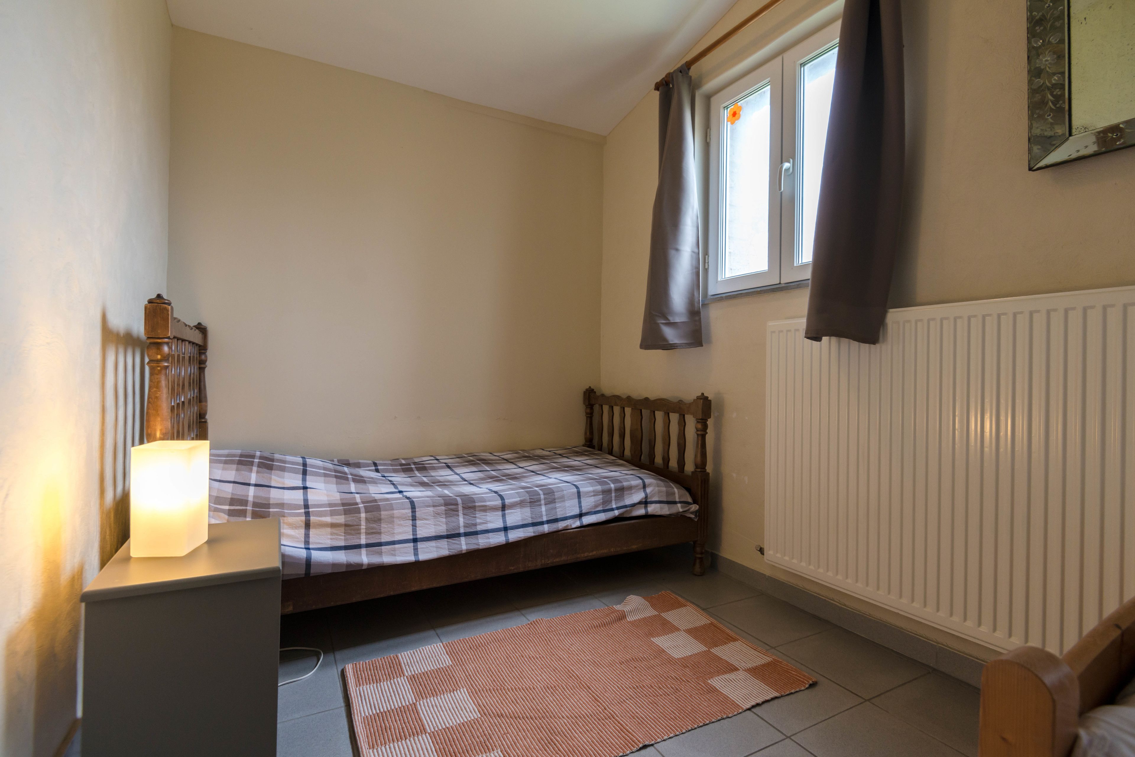 Una habitación pequeña con una cama individual, cabecera de madera, mesita de noche con lámpara, ventana con cortinas y radiador.