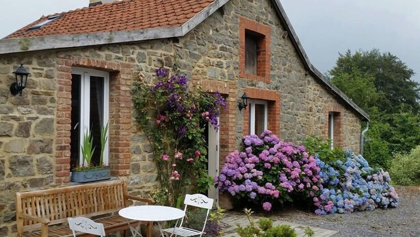 A stone house with a sloped roof, a small porch with a bench and table, and a garden with hydrangea bushes. - Gîte Les Trois Voisins (Gedinne)