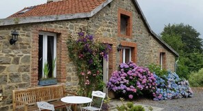 Ein Steinhäuschen mit einem geneigten Dach, einer kleinen Veranda mit Bank und Tisch sowie einem Garten mit Hortensienbüschen.