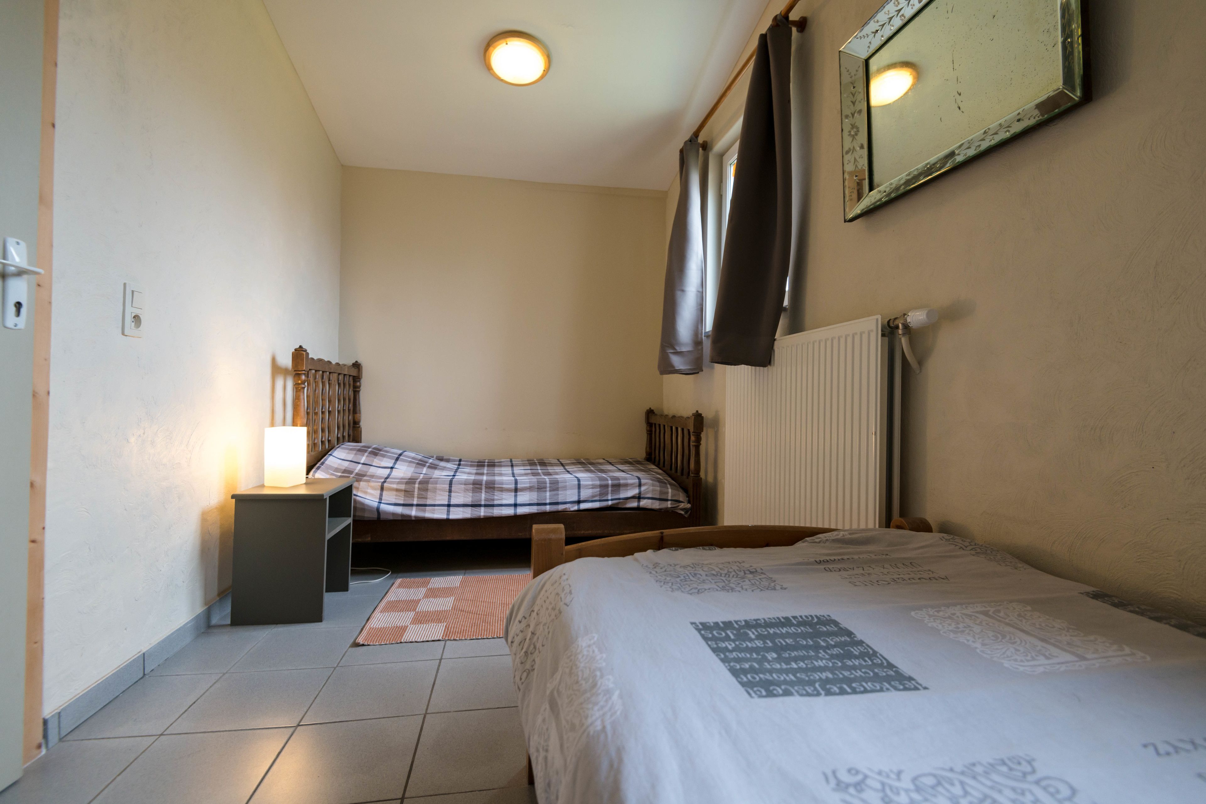 Een kleine, schone kamer met twee bedden, een nachtkastje, een radiator en een spiegel aan de muur.