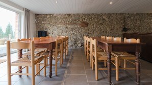 A dining area with a stone wall, wooden tables and chairs, and a view of the outdoors. - Gîte Les Trois Voisins (Gedinne)