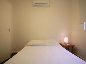 A small, simple room with a bed, a bedside table with a lamp, and a wall-mounted air conditioning unit.