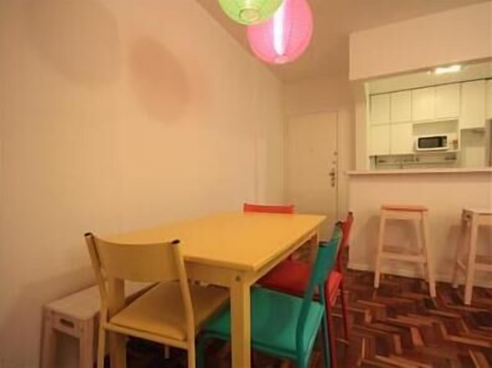 Un petit coin repas avec une table et des chaises jaunes, un parquet à motifs en chevrons et une cuisine visible à travers une fenêtre.