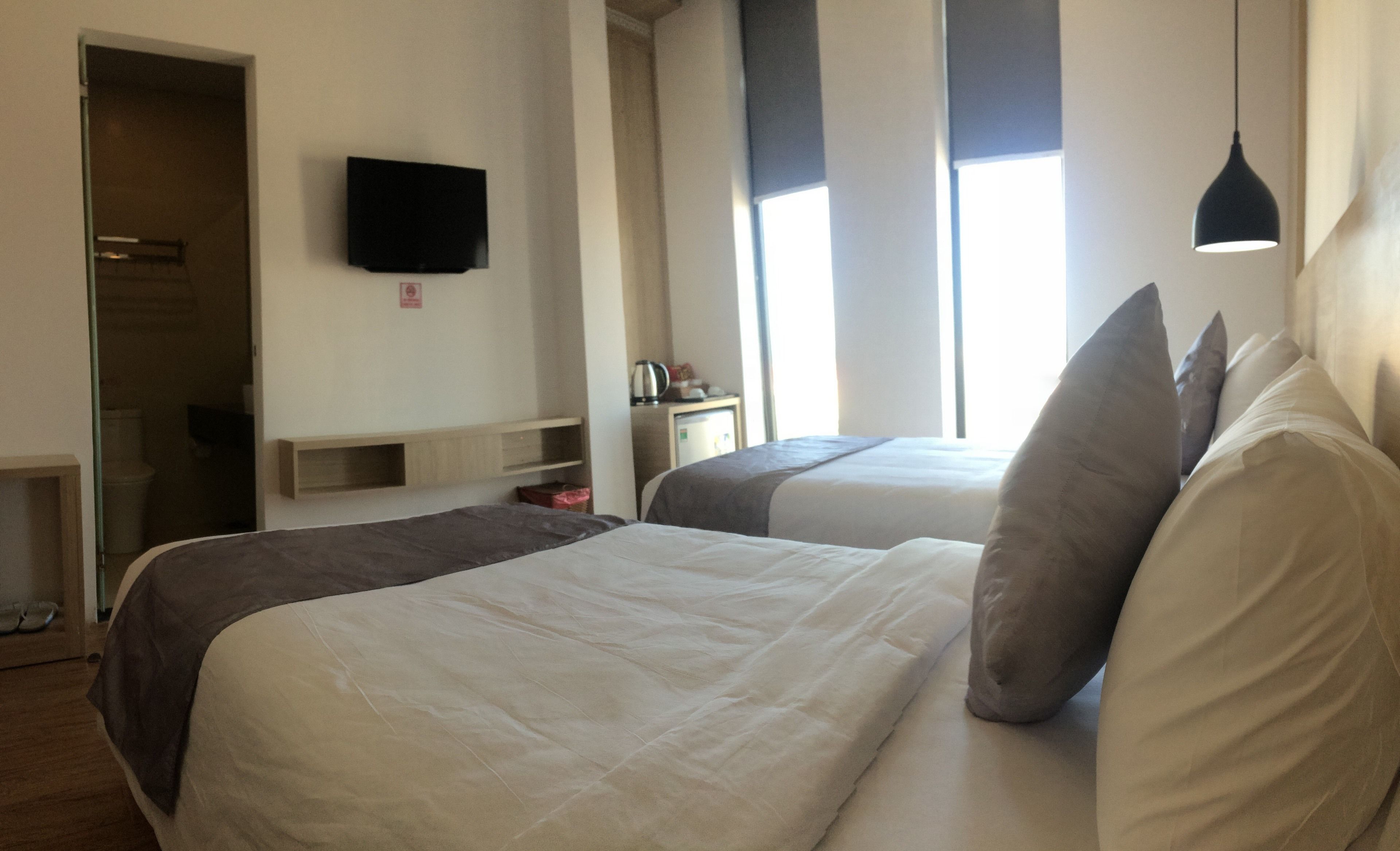 Ein Hotelzimmer mit zwei Betten, einem an der Wand befestigten Fernseher, einem Schreibtisch mit Wasserkocher und einem Fenster, das für natürliches Licht sorgt.
