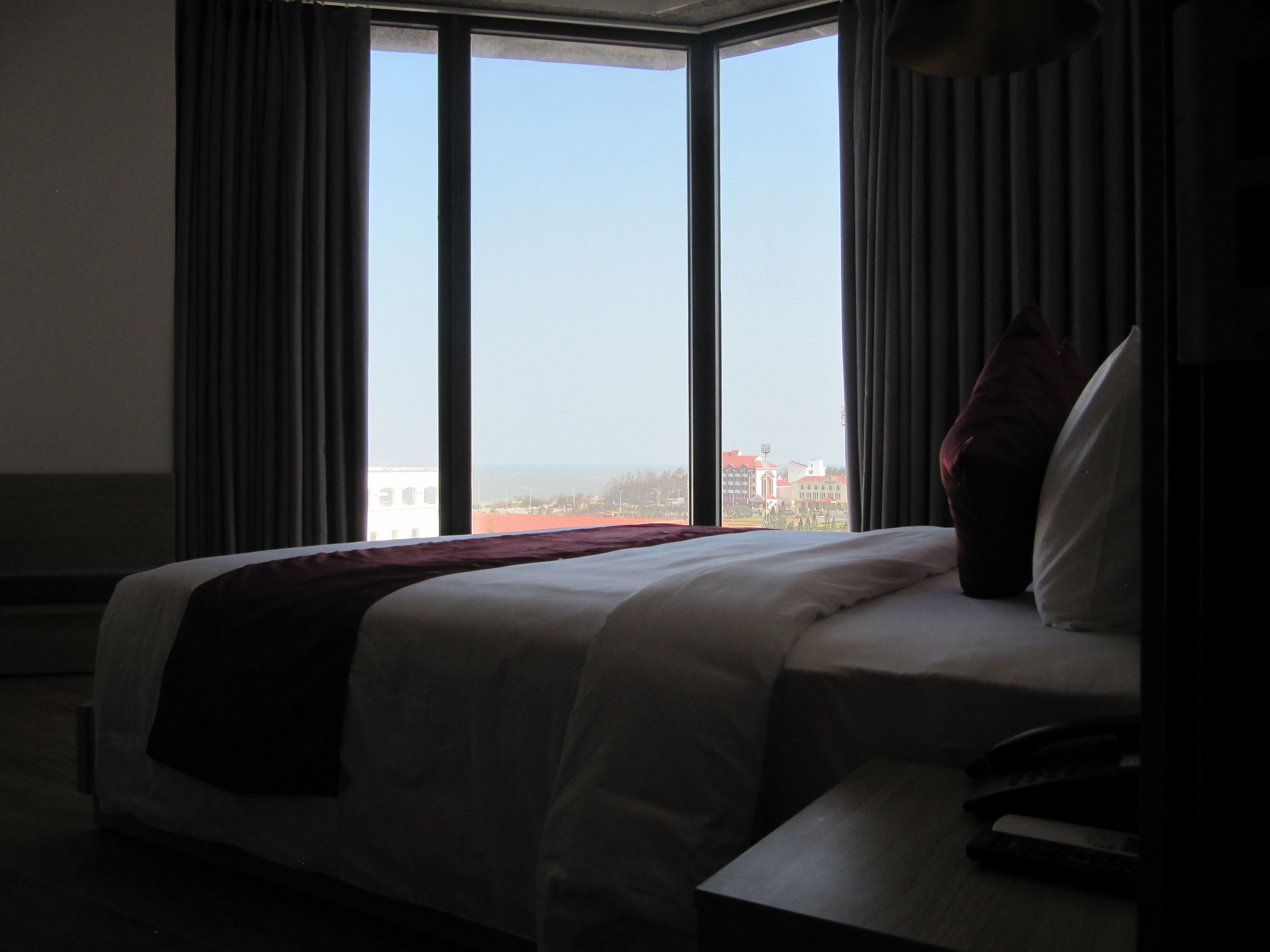 A hotel room with a large bed, a view of the cityscape through floor-to-ceiling windows, and a remote control on the bedside table.