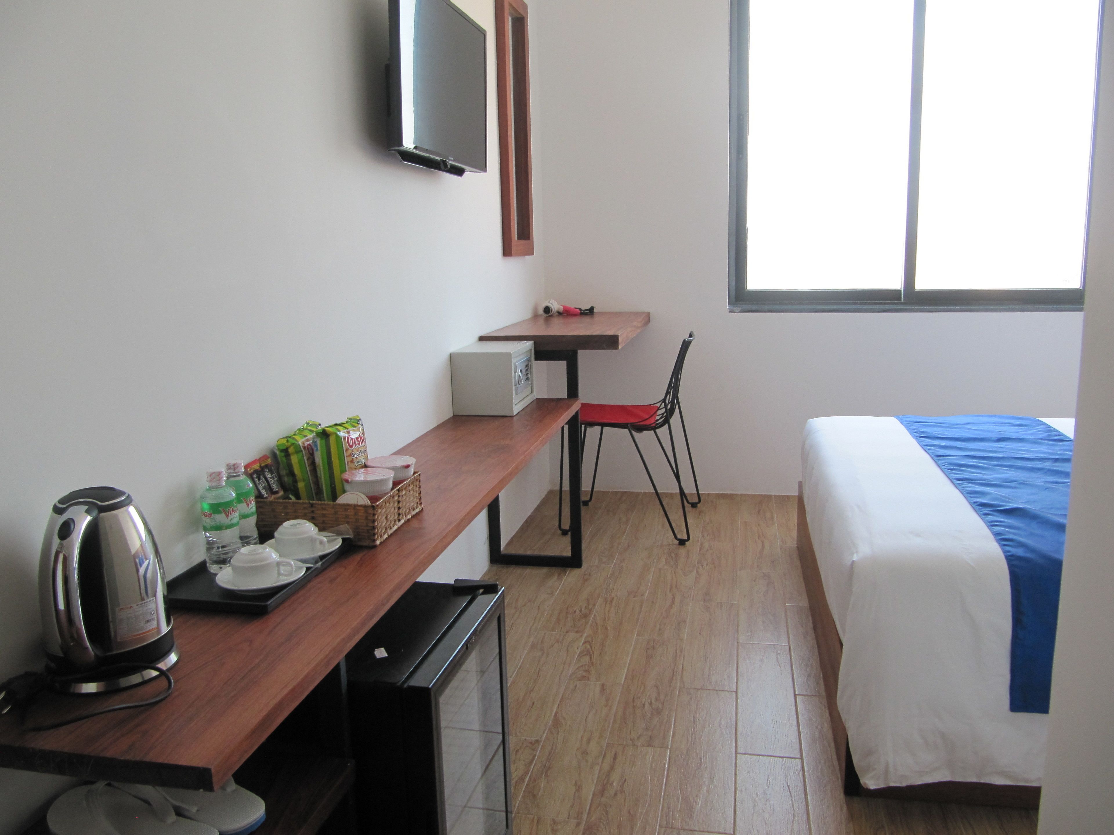 A hotel room with a bed, a desk with a kettle, a chair, a TV, and a window.