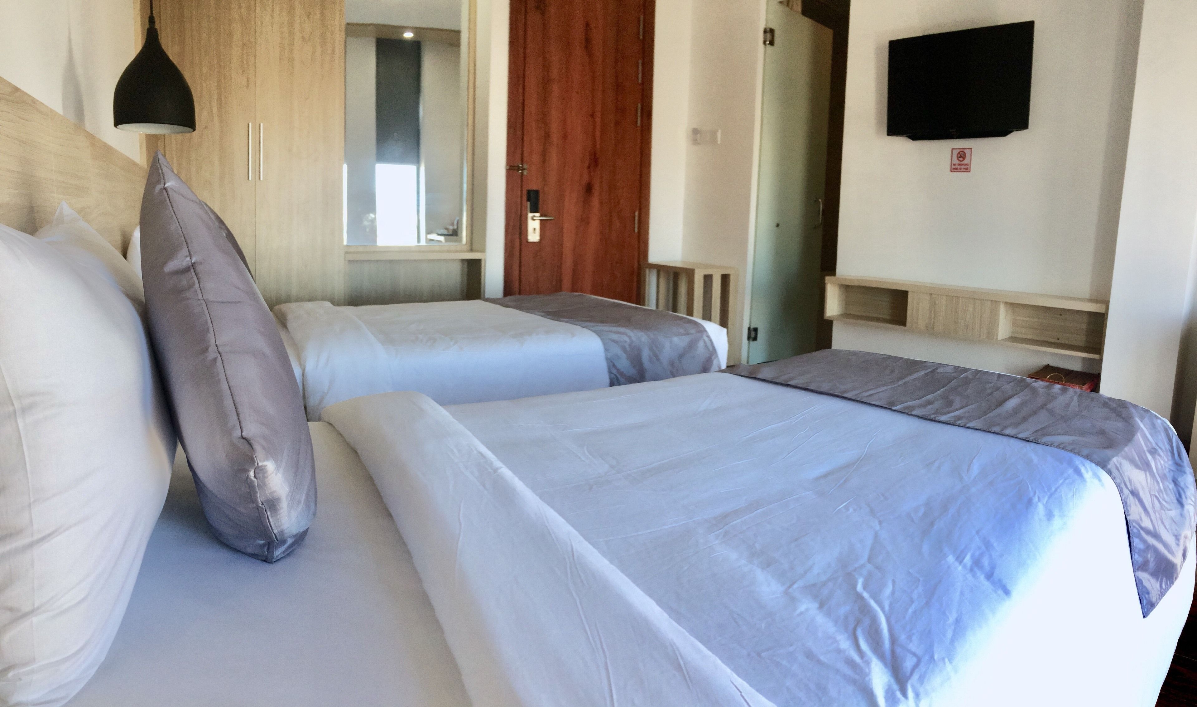 A hotel room with two beds, a wooden headboard, a mirror, a television on the wall, and a door leading to another room.
