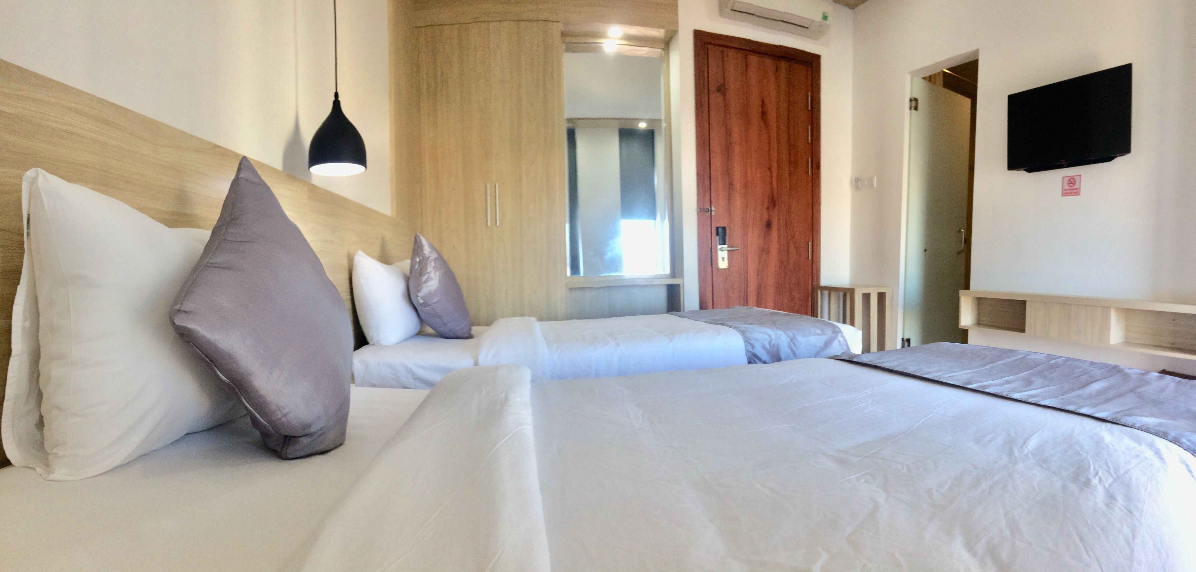 Habitación de hotel con dos camas, cabecera de madera, lámpara colgante negra, armario de madera, televisor montado en la pared y una puerta que da a otra habitación.