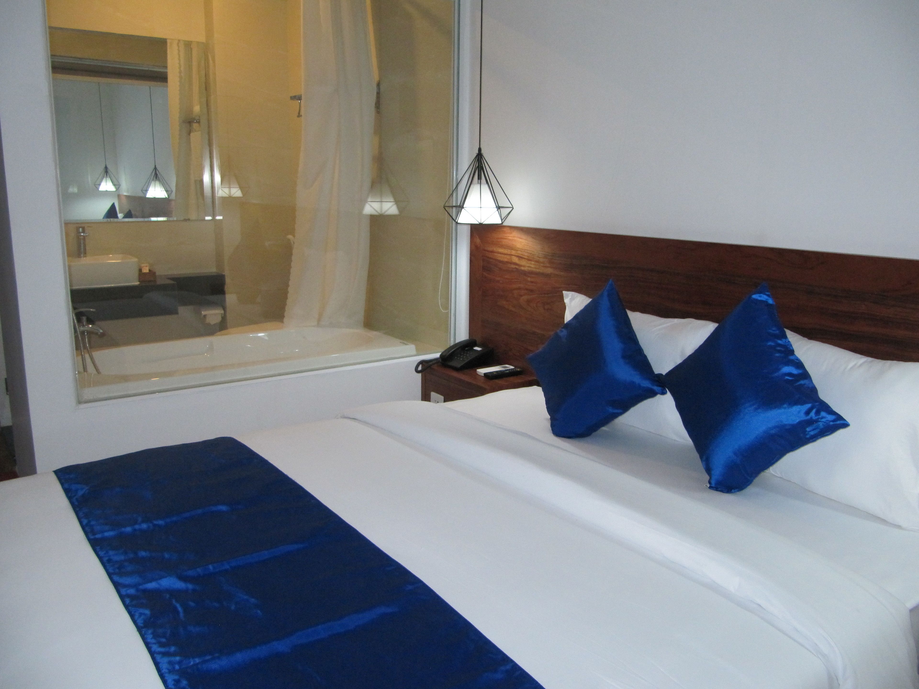 A hotel room with a large bed, a wooden headboard, two blue pillows, a bedside table with a phone and remote control, and a window with a view of a bathroom and a mirror.