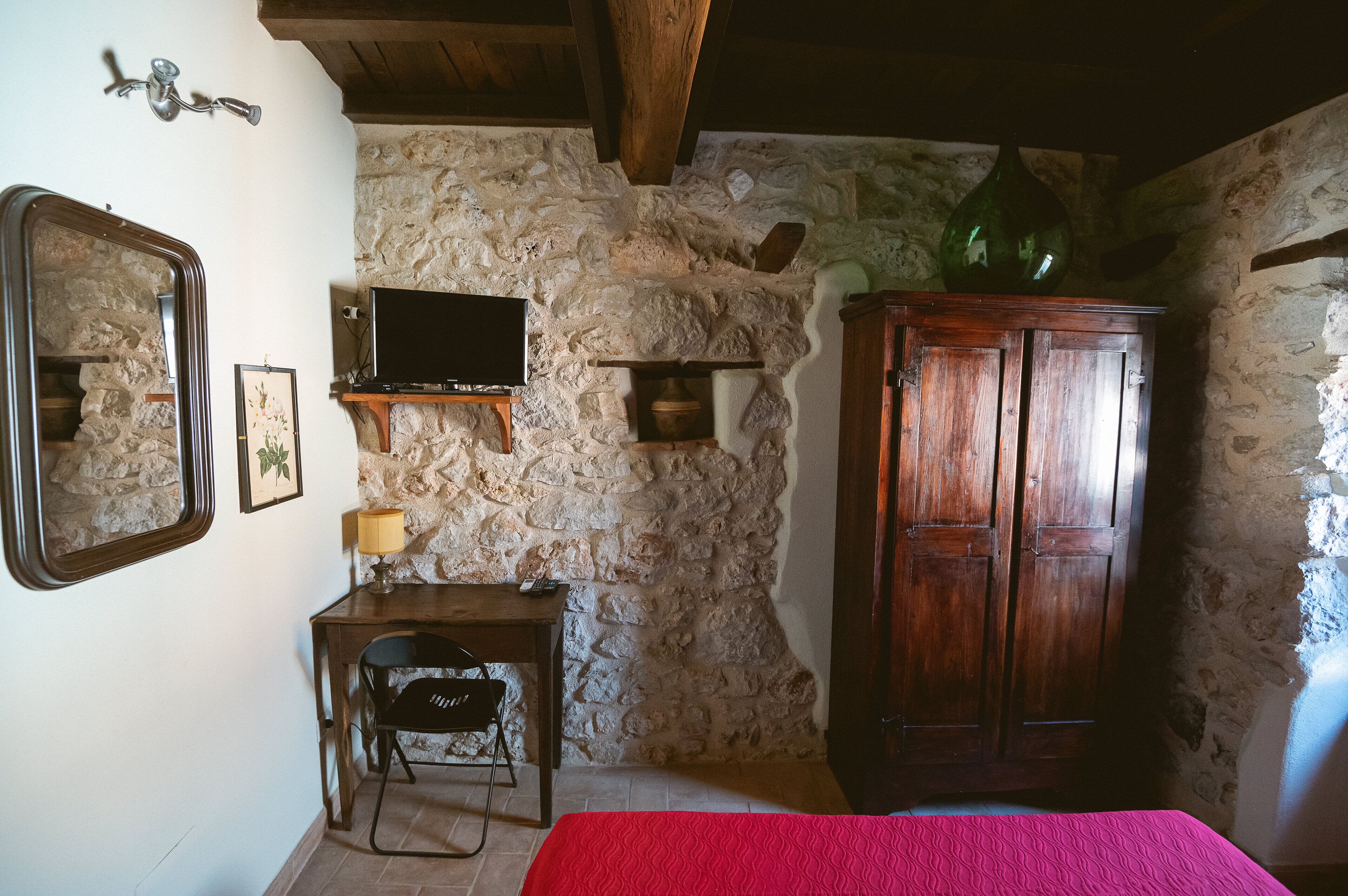 A rustic room with stone walls, a wooden cabinet, a desk with a chair, a television, and a bed with a red bedspread.