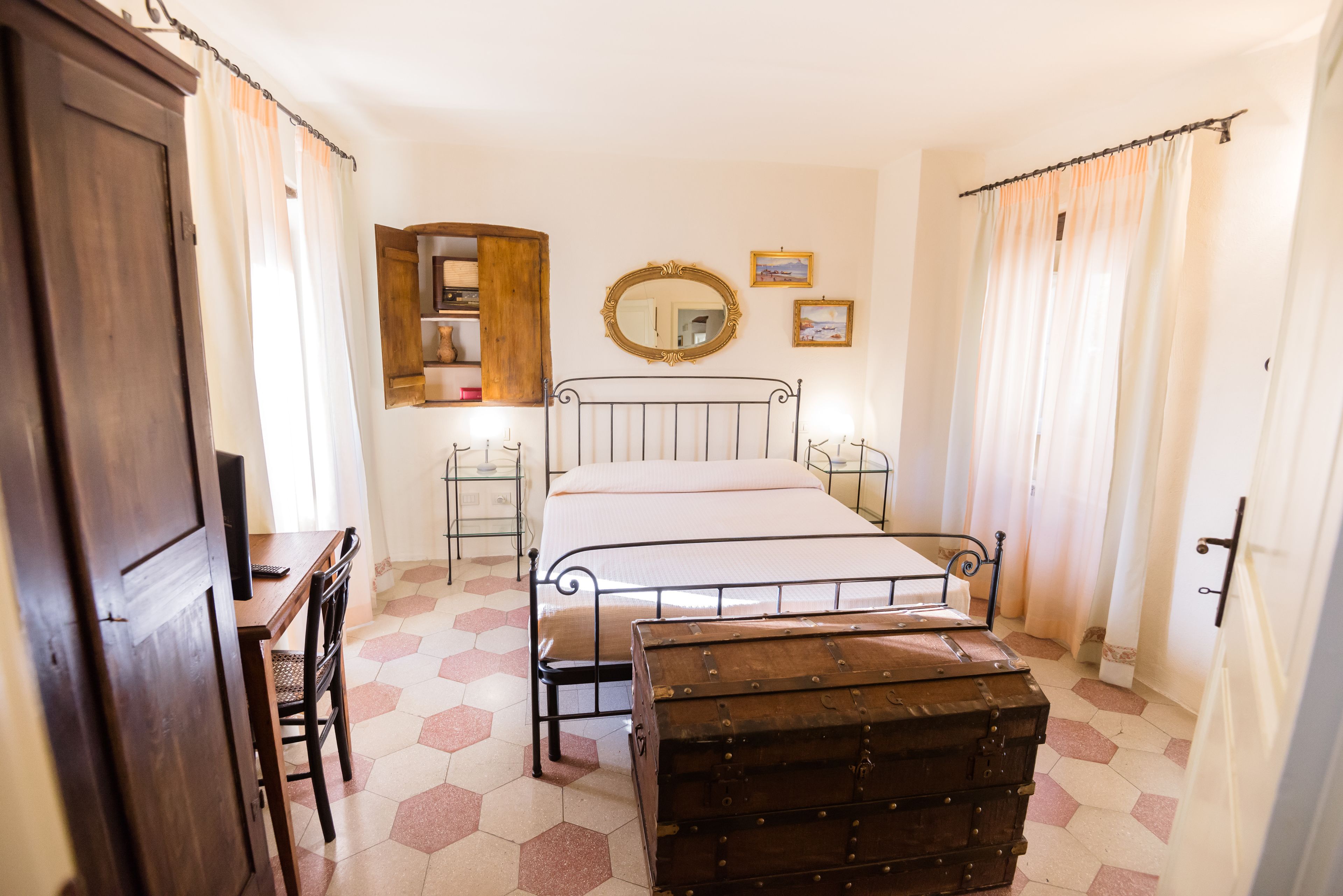 A bedroom with a bed, a wooden chest, a desk with a chair, a mirror, and a shelf with decorative items.
