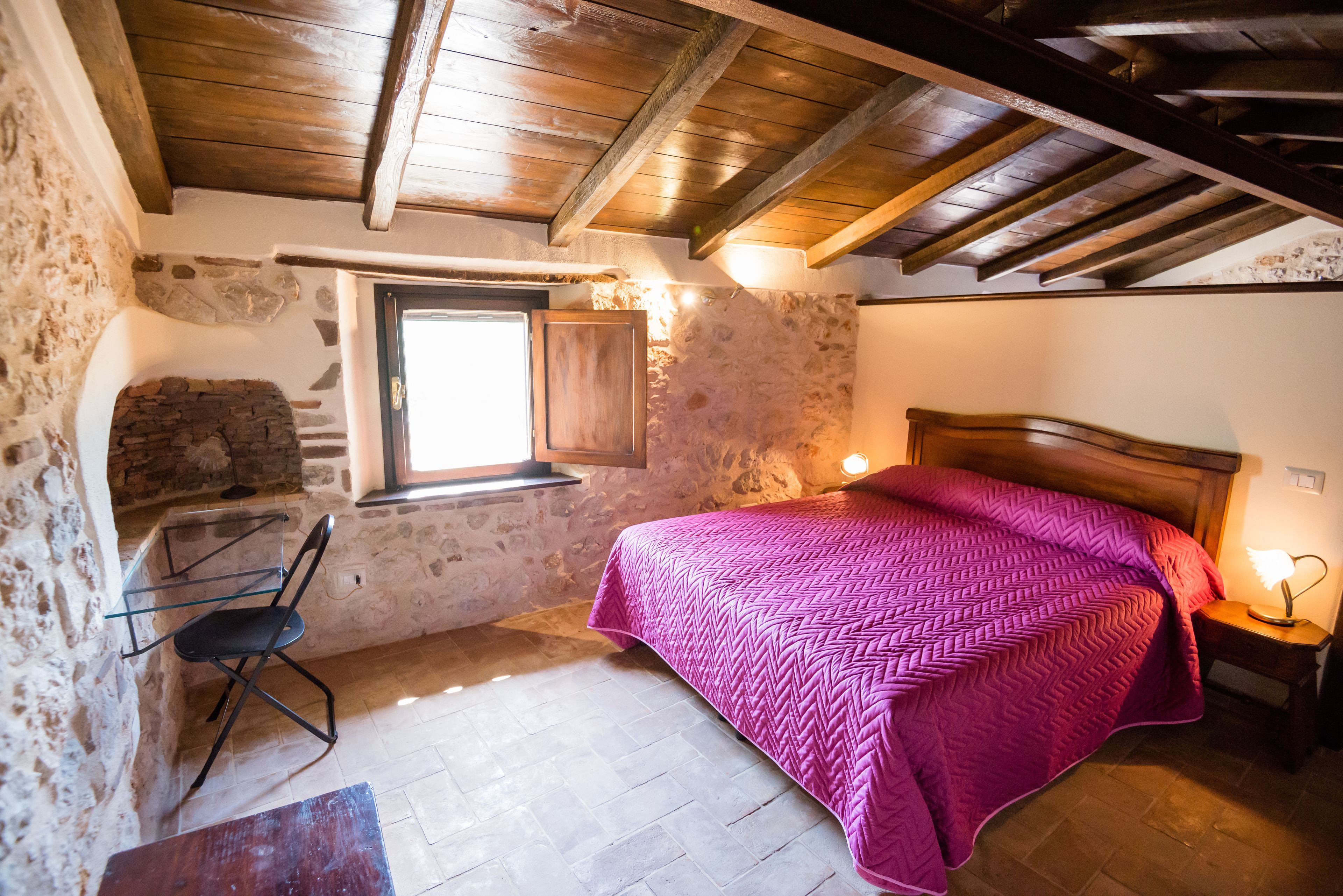 A bedroom with a stone wall, wooden beams, a bed with a pink quilt, a desk with a chair, and a window.