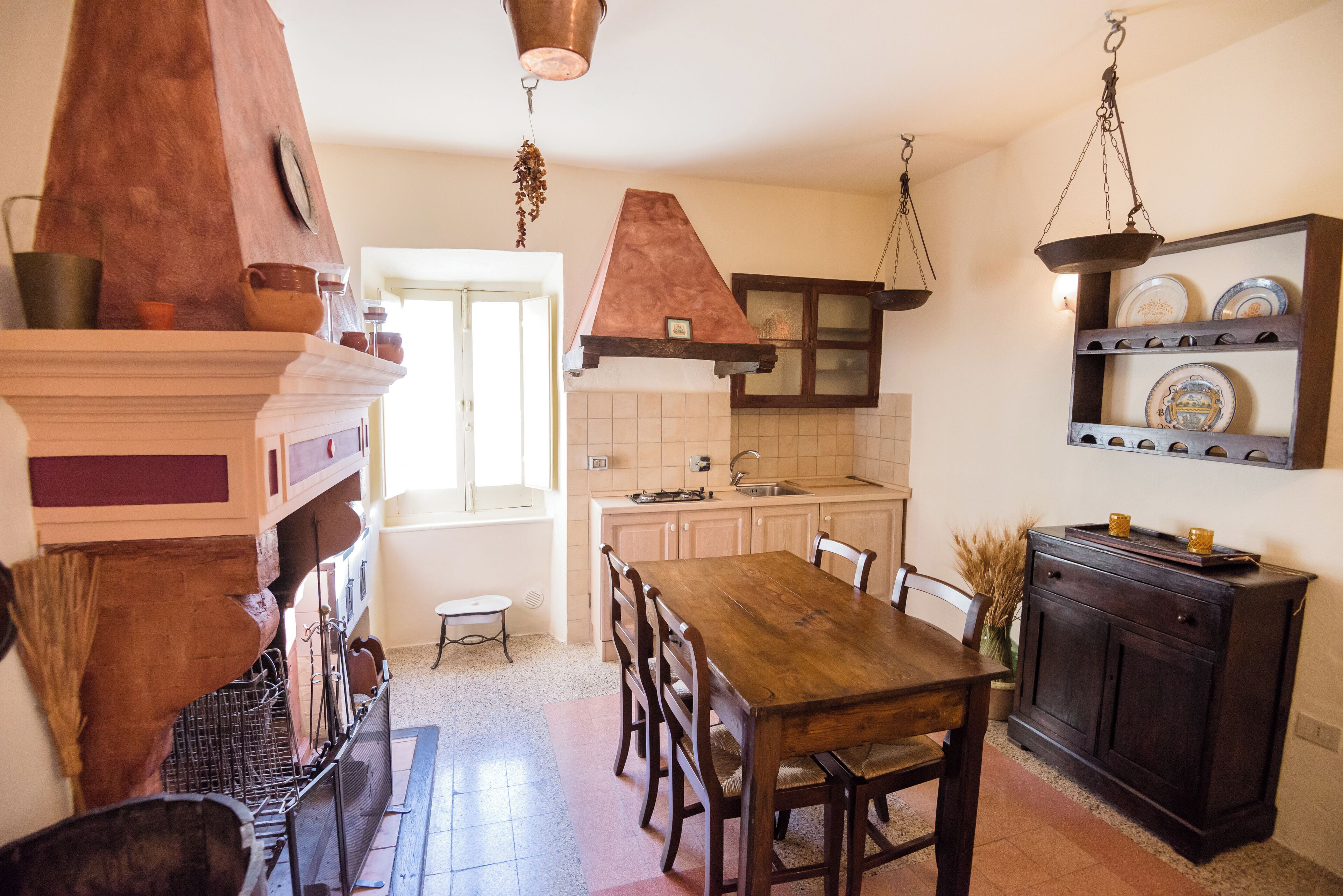 Una cocina con una mesa de madera, sillas, un colgador para ollas y una repisa con vajilla.