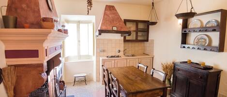 Una cocina con una mesa de madera, sillas, un colgador para ollas y una repisa con vajilla.