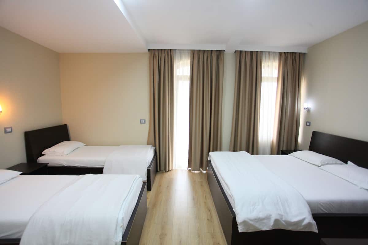 A hotel room with three single beds, a wooden floor, beige walls, and a window with curtains.