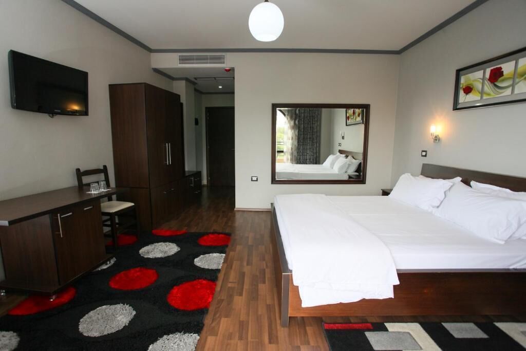 A hotel room with a bed, a desk, a chair, a TV, a mirror, and a red and black patterned carpet.