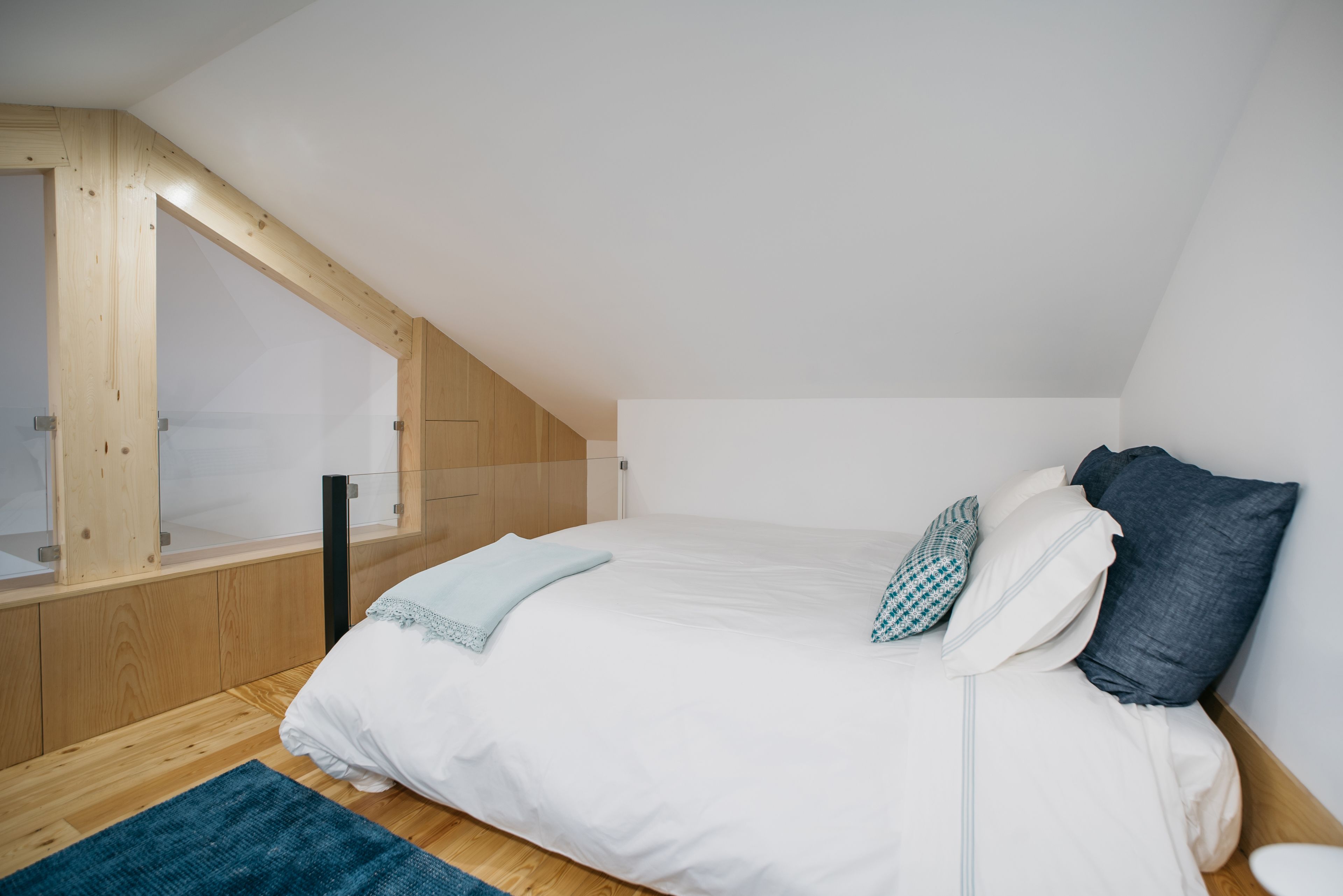 Een slaapkamer met een schuine kap, een bed met wit beddengoed, een blauwe plaid en een houten hoofdbord.