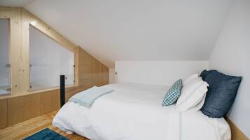 Een slaapkamer met een schuine kap, een bed met wit beddengoed, een blauwe plaid en een houten hoofdbord.