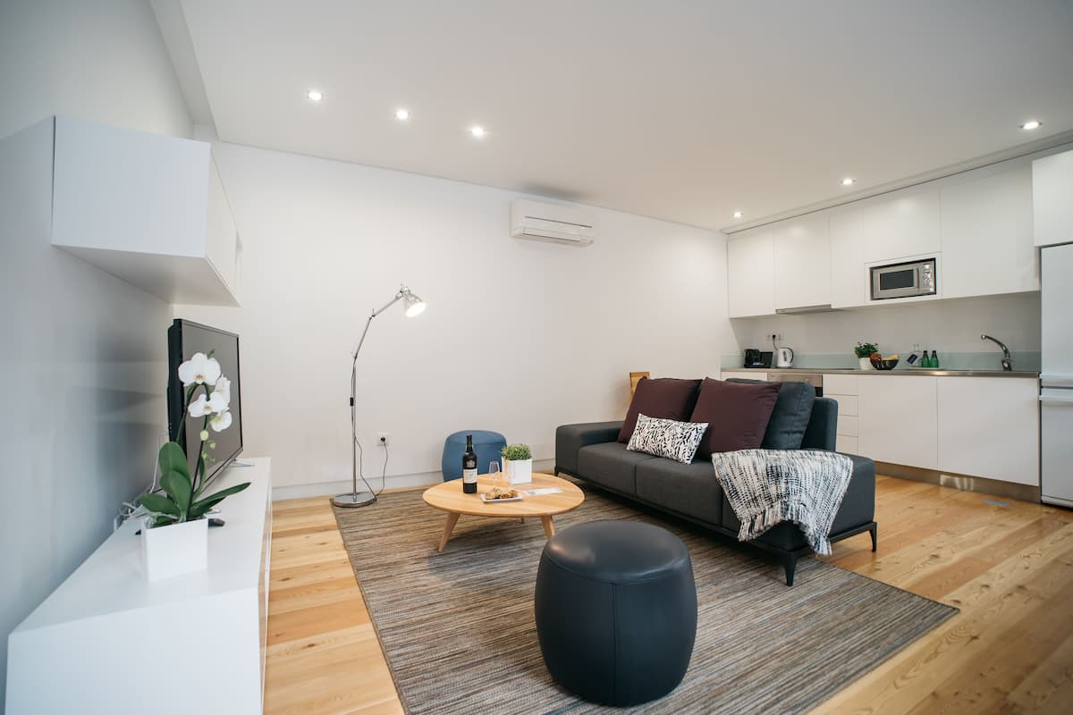 A modern living room with a sofa, coffee table, and ottoman. There is a kitchen area with a microwave and cabinets.