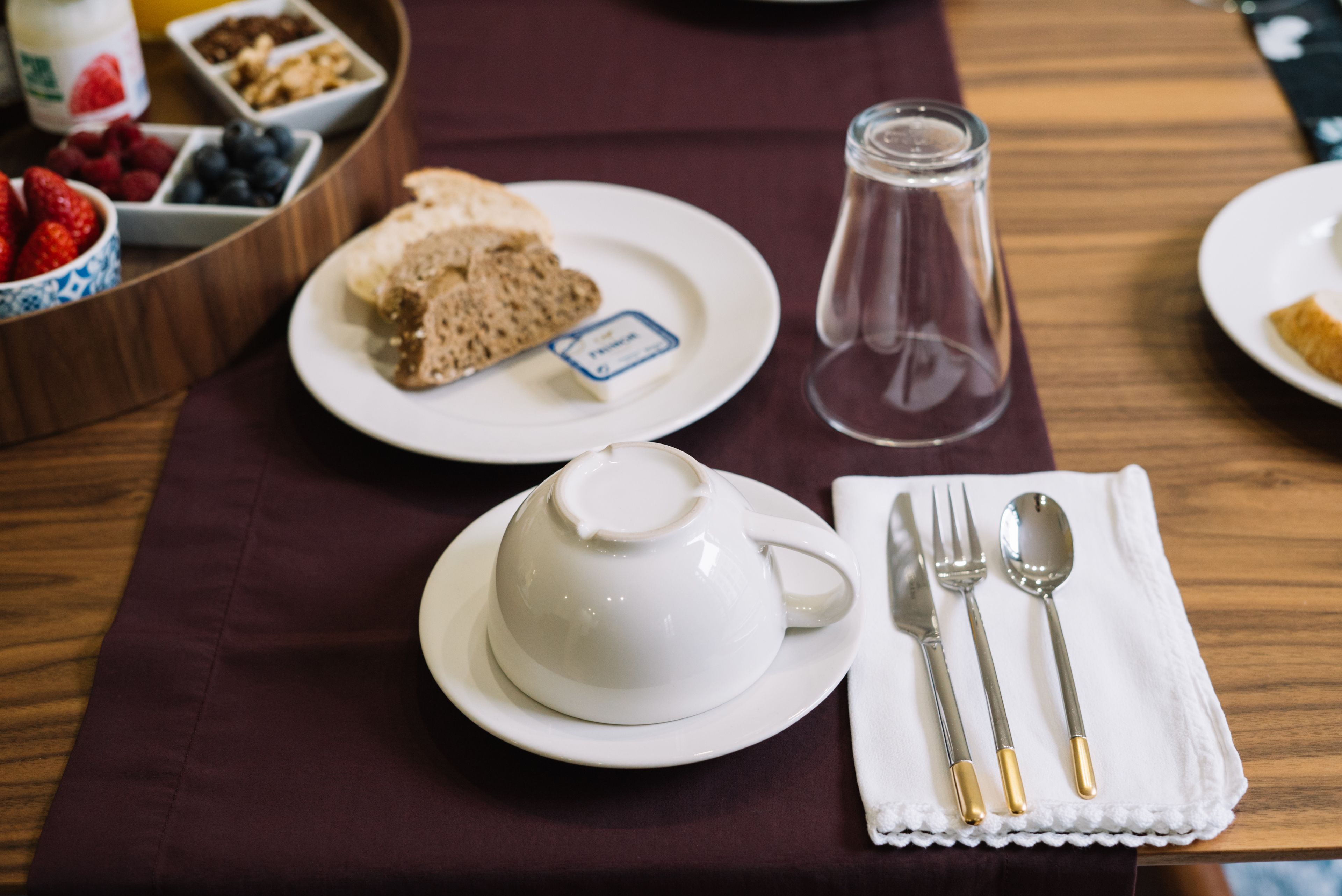 Una mesa dispuesta con tetera, un plato de pan y una etiqueta, una jofaina de vidrio y una servilleta con cubiertos.