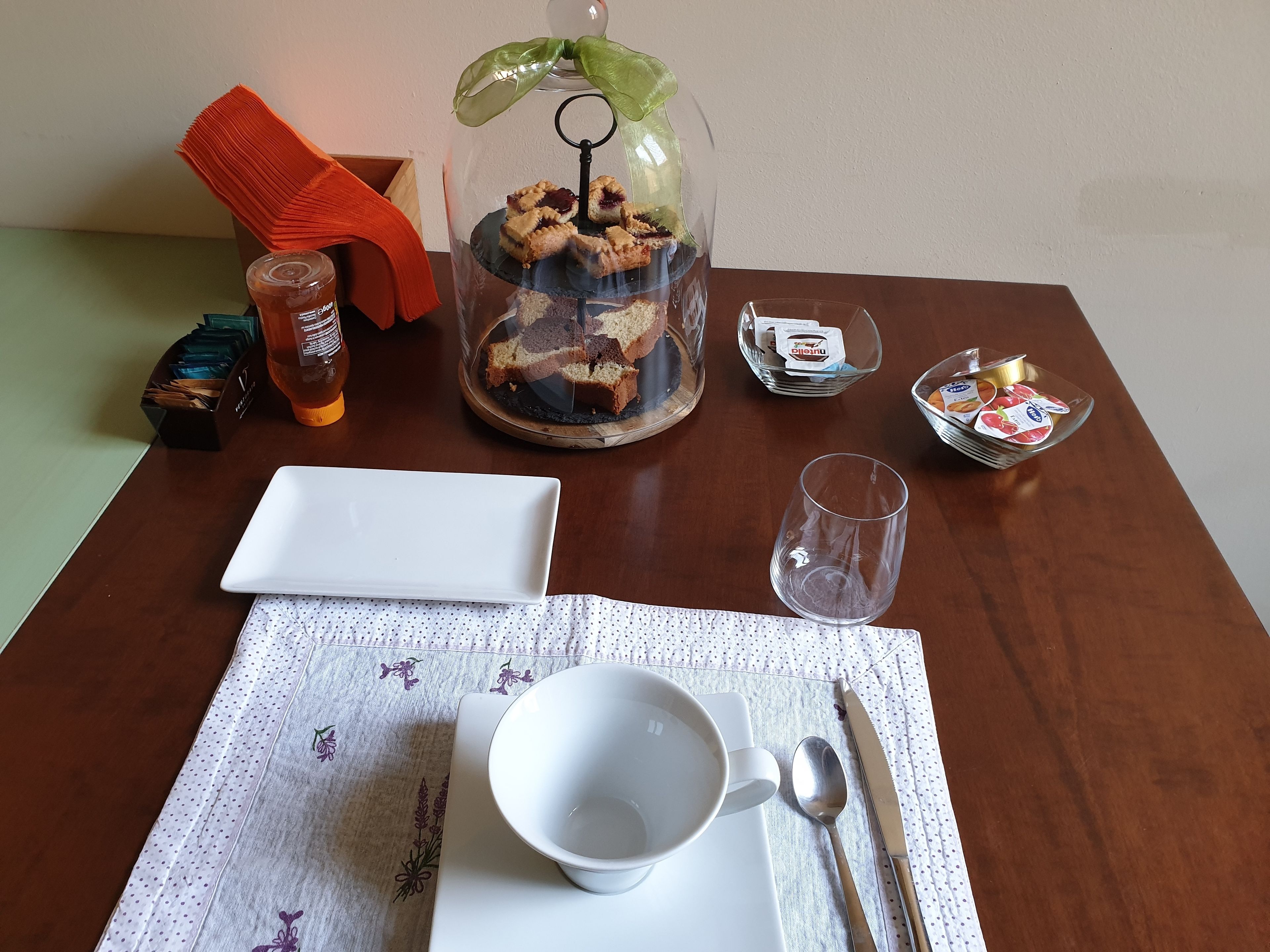 A neatly arranged table with a glass dome containing pastries, a white plate, a glass, a spoon, and a napkin.