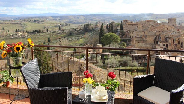 A balcony with wicker chairs, a table with a vase of flowers, and a view of a rural landscape with buildings. - B&B Ridolfi (San Gimignano)