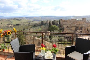 Un balcon avec des fauteuils en osier, une table ornée d’un vase de fleurs et une vue sur un paysage rural avec des bâtiments.