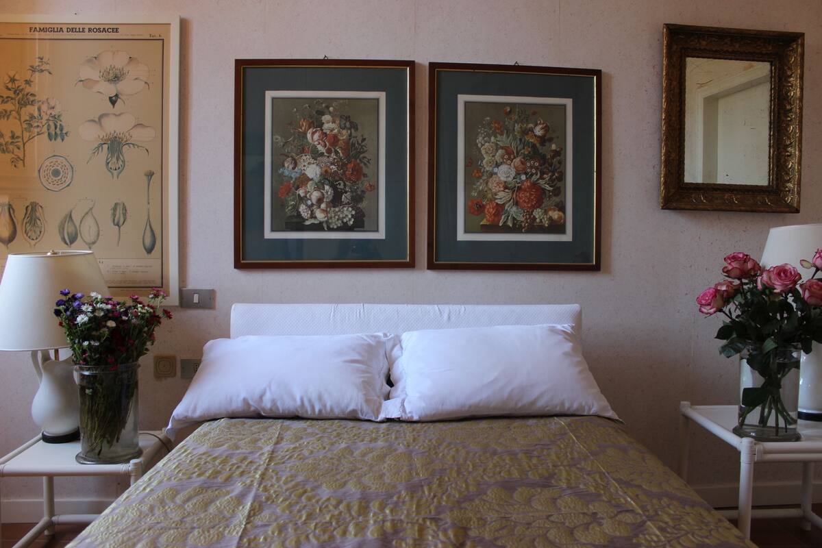 A neatly made bed with white pillows, a floral-patterned bedspread, and two framed floral paintings on the wall.