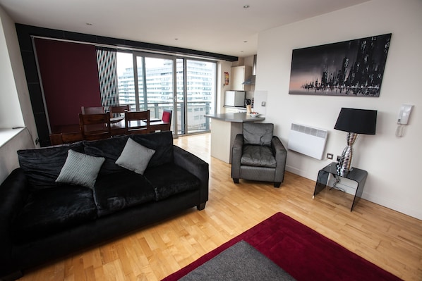 Ein modernes Wohnzimmer mit einer schwarzen Couch, einem gestreiften Sessel und einem roten Teppich. An der Wand hängt ein Stadtbild, und durch die Glastür ist eine Küchenzeile erkennbar.