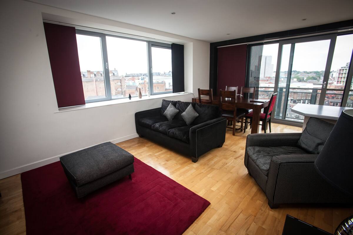 A modern living room with a sofa, ottoman, and a dining area with chairs and a table. Large windows offer a city view.