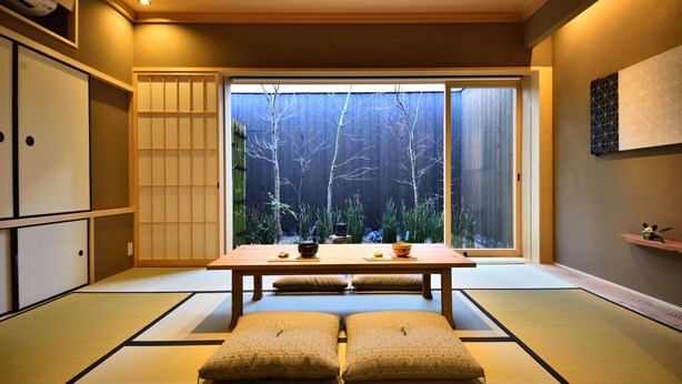 Ett traditionellt japanskt rum med tatamimattor på golvet, ett lågt bord med kuddar, skjutdörrar och utsikt över en trädgård.