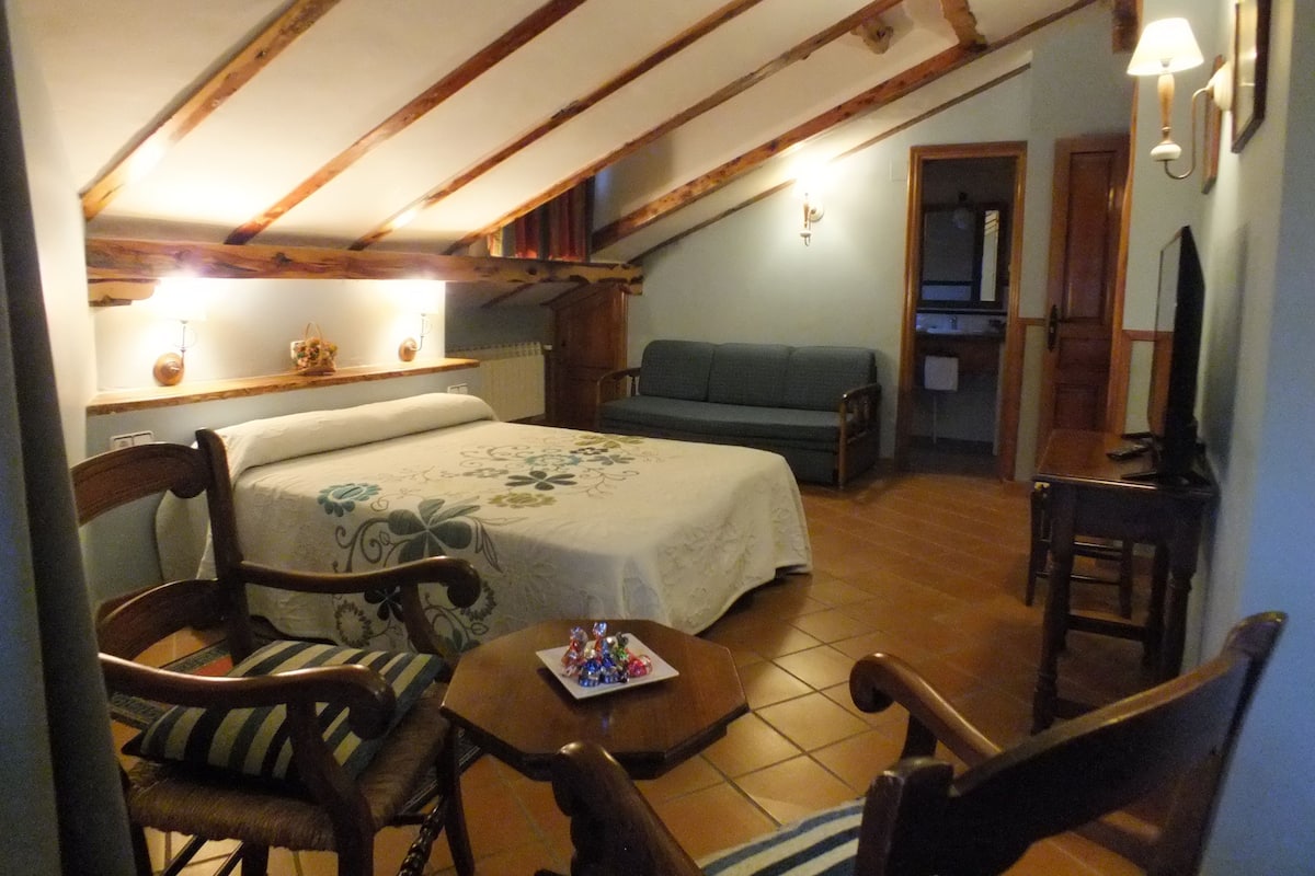 A cozy room with a bed, a sofa, and a small table. There is a wooden chair and a lamp on the wall.