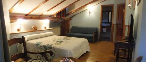 Una habitación acogedora con una cama, un sofá y una pequeña mesa. Hay una silla de madera y una lámpara en la pared.