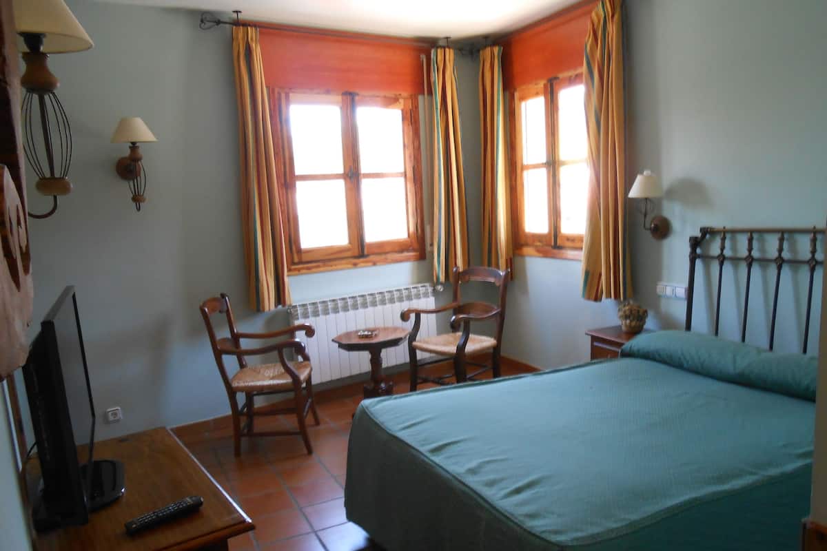 A hotel room with a bed, a desk with a TV and remote, two chairs, a small table, and windows with curtains.
