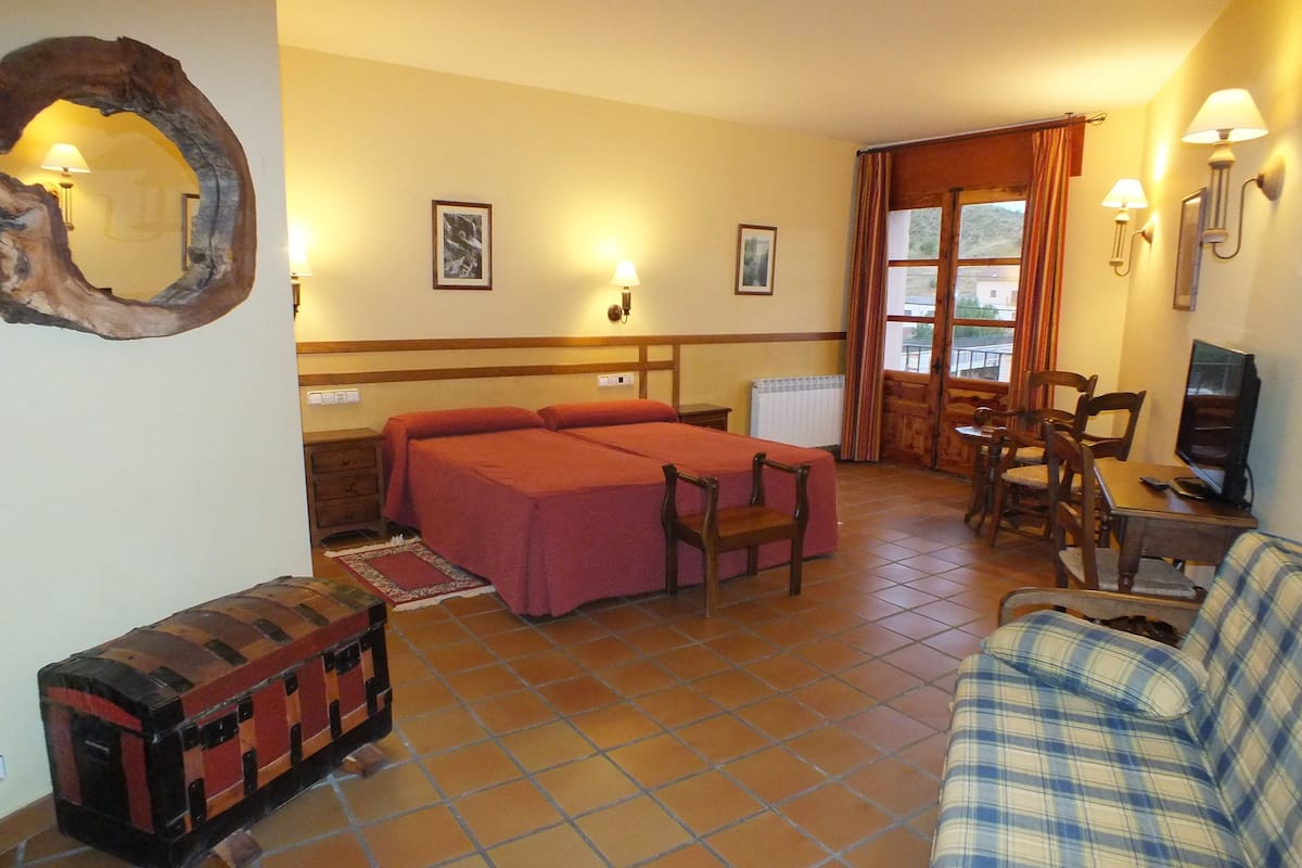 A hotel room with two beds, a sofa, a small table, a TV, and a window with curtains.