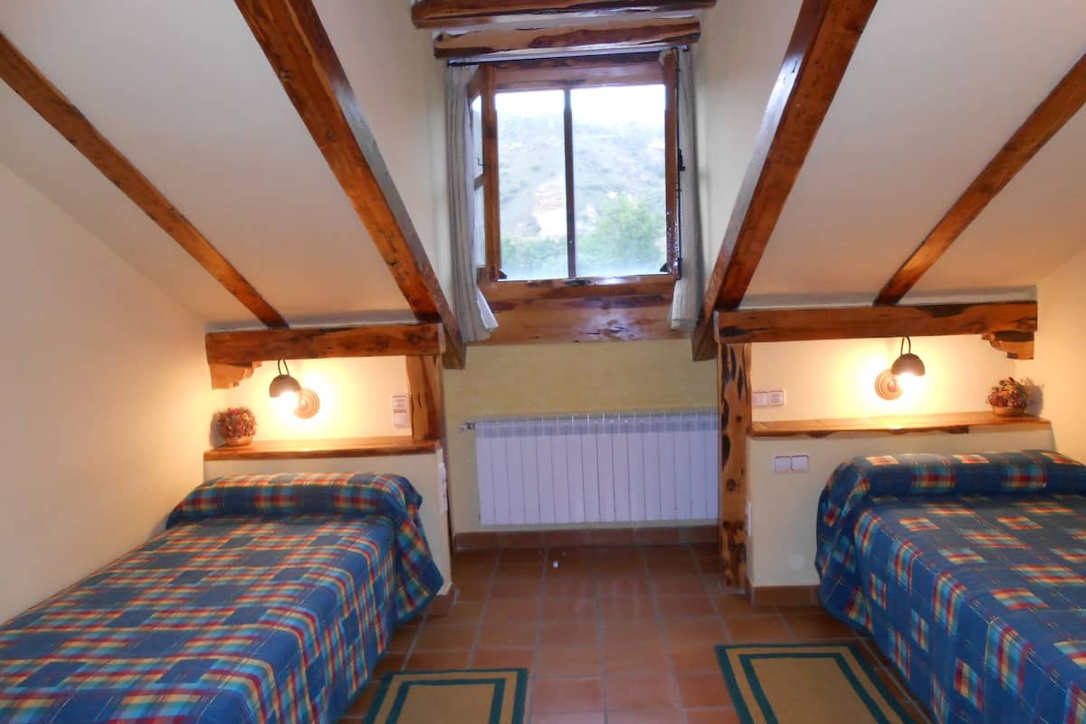 A room with two beds, wooden beams, a window, a radiator, and a shelf with a lamp.