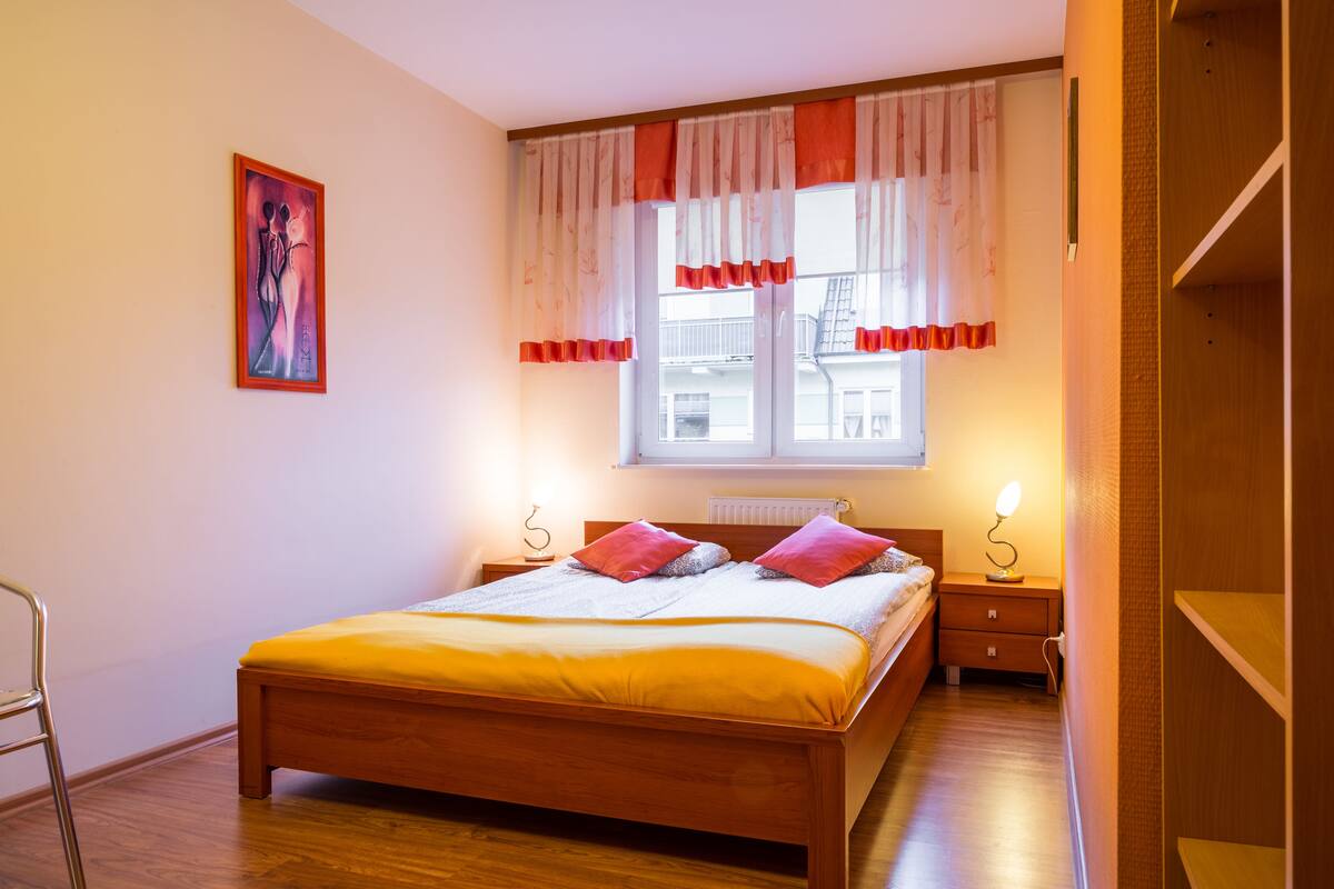 A bedroom with a wooden bed, two bedside tables with lamps, a window with curtains, and a painting on the wall.