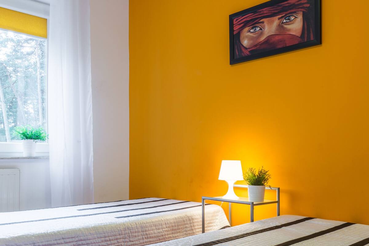 A bedroom with two beds, a window with white curtains, a bedside table with a lamp and a plant, and a painting on the wall.