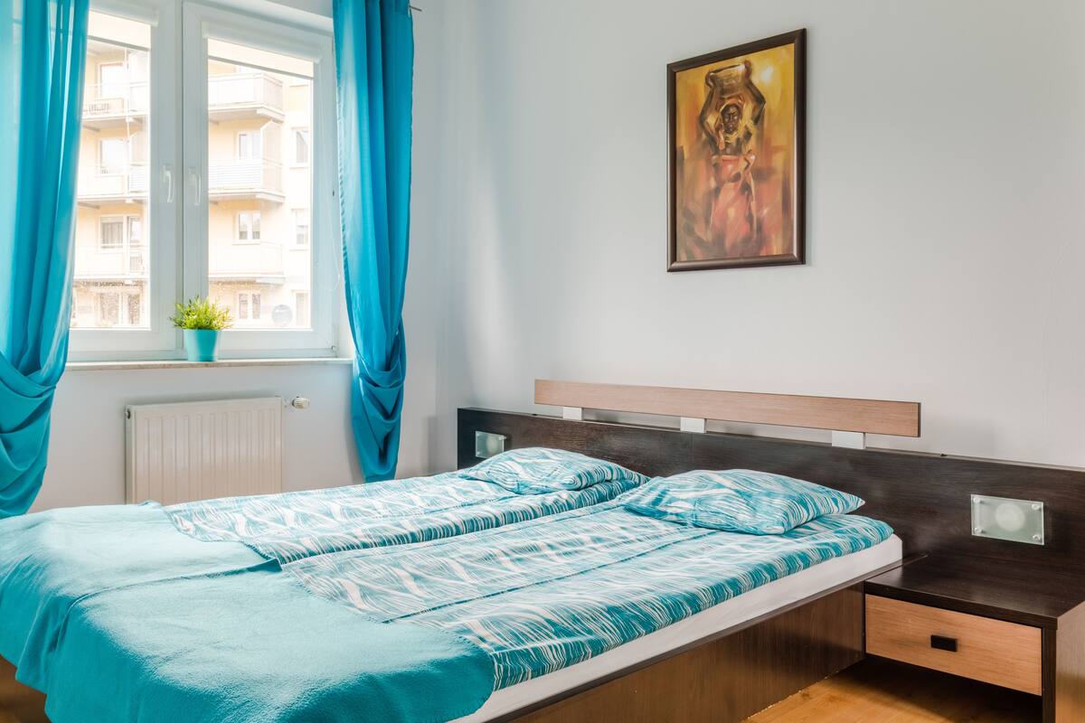 A bedroom with a bed, a window with curtains, a radiator, a potted plant, and a painting on the wall.