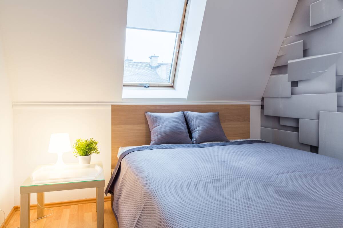 A modern bedroom with a skylight, a bedside table with a lamp and plant, and a bed with a grey bedspread.