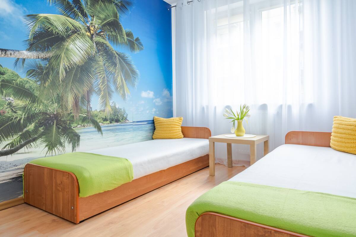 A room with two beds, a mural of a beach, and a small table with a vase.