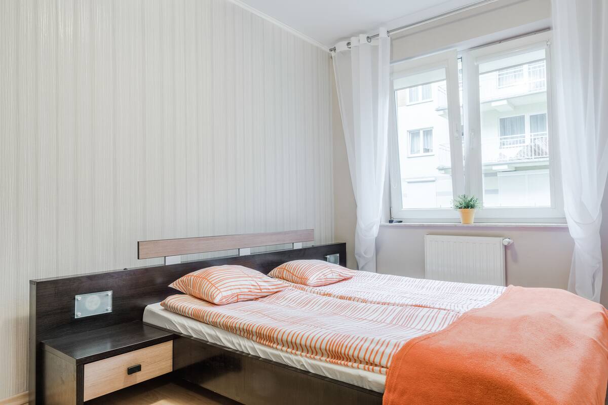 A neatly made bed with striped bedding, a bedside table with a drawer, and a window with curtains.