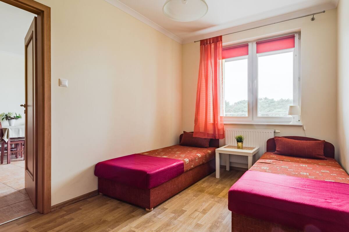 A small, well-lit room with two single beds, a small table, and a window with a view.