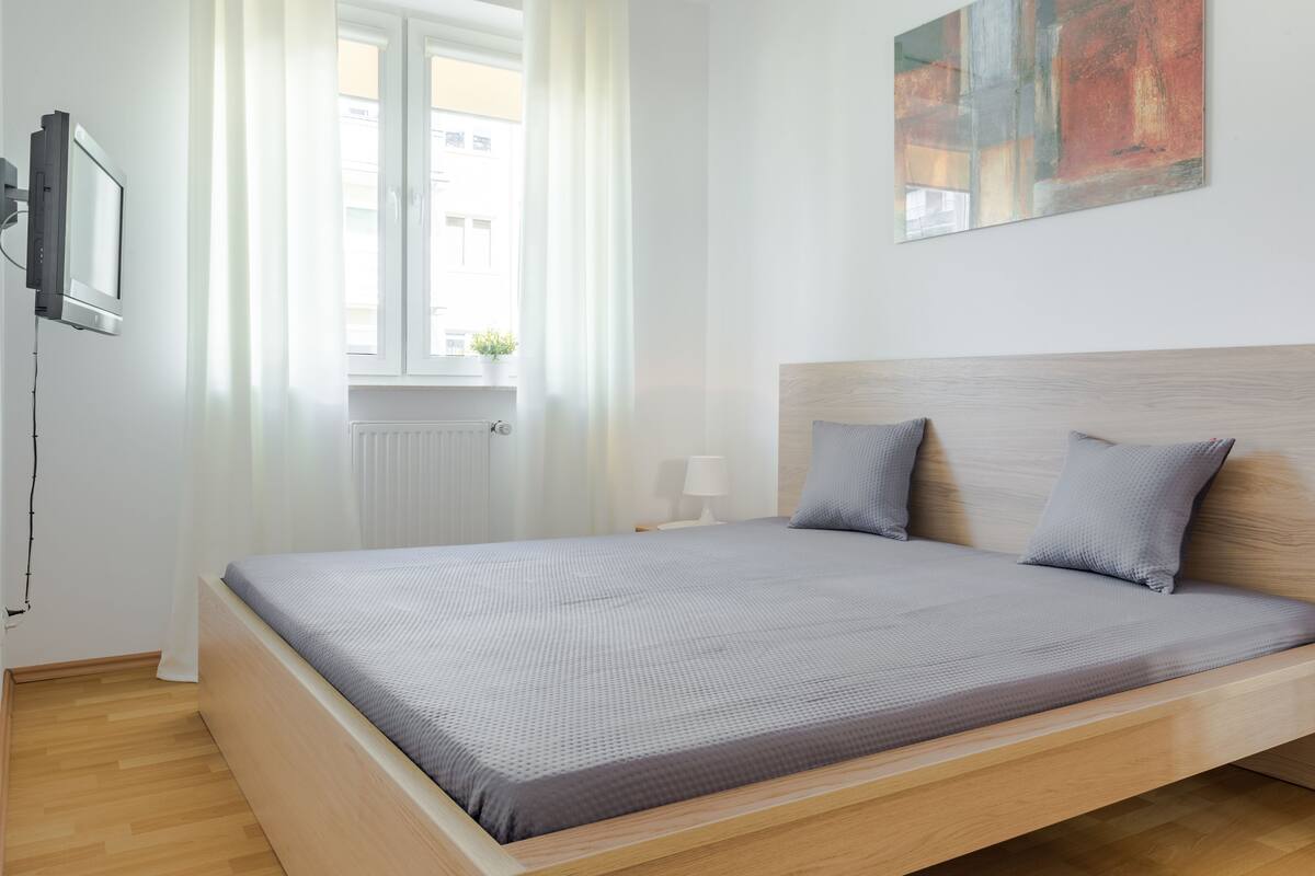 A modern bedroom with a wooden bed frame, a grey bedspread, and a window with sheer curtains.