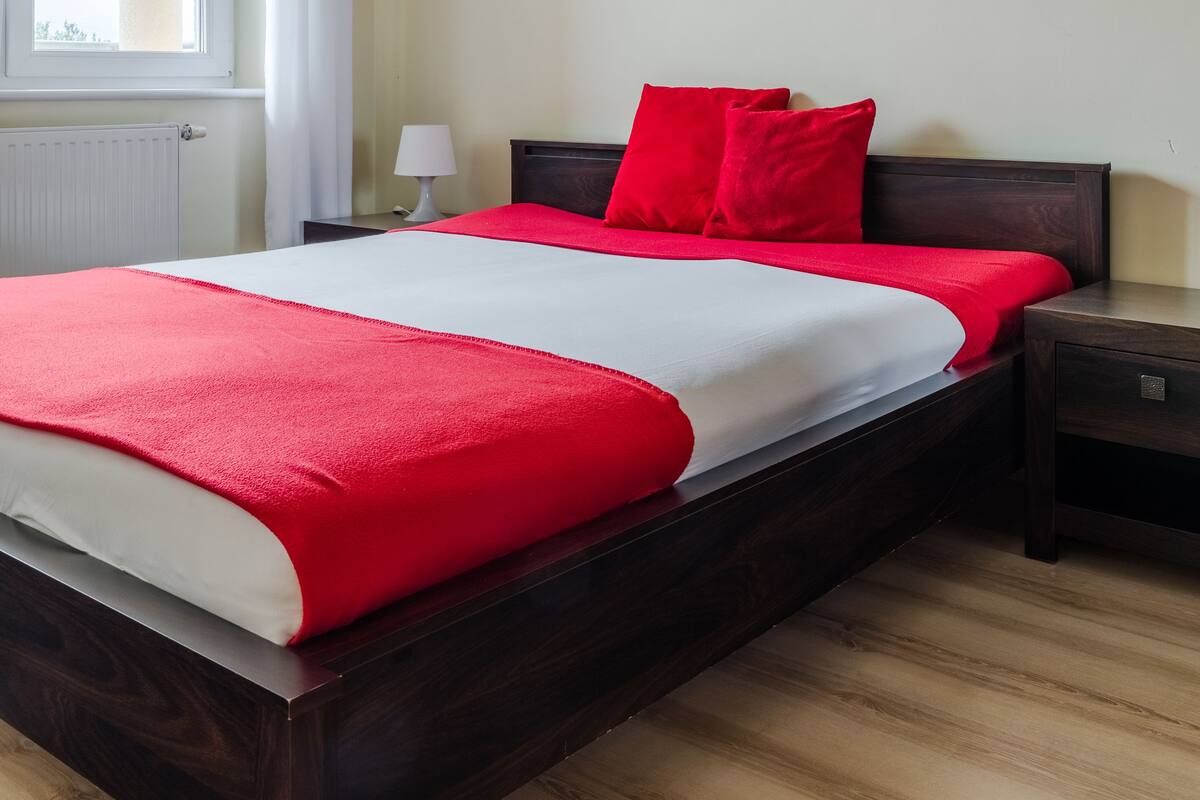A bedroom with a wooden bed, red and white bedding, a nightstand, a lamp, and a window with curtains.