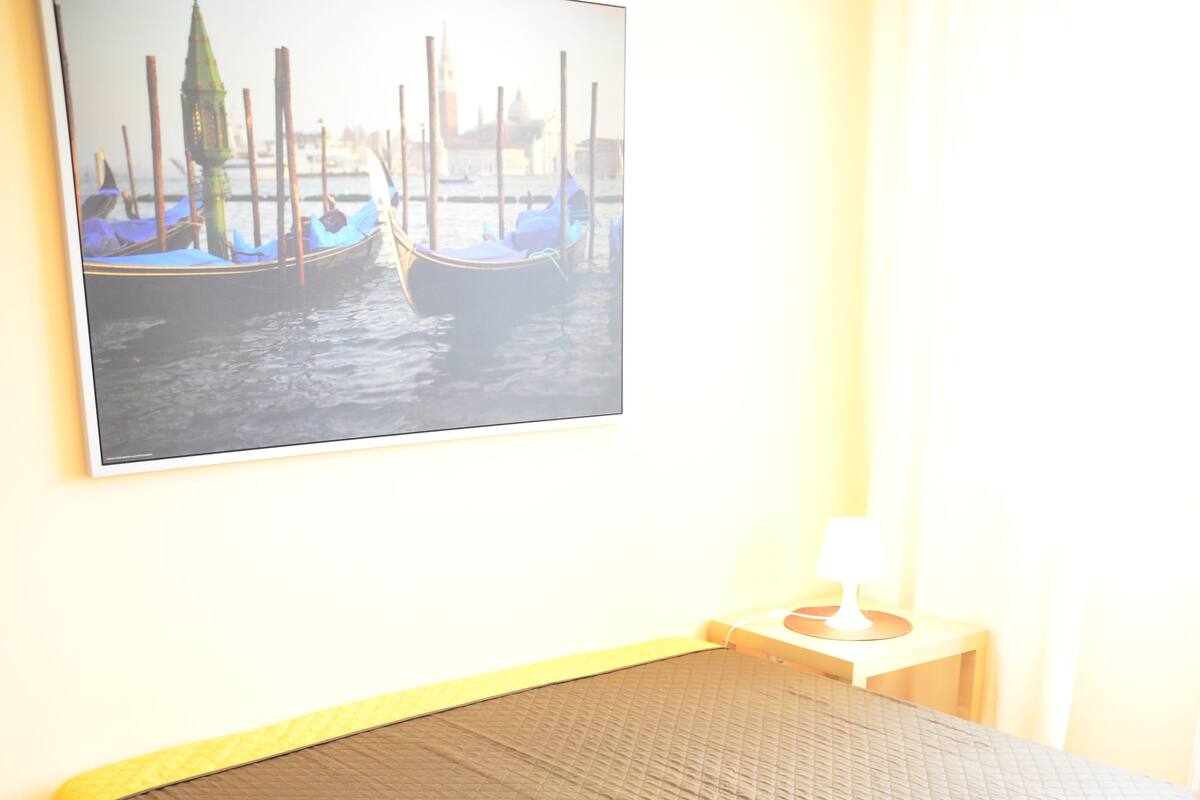 A bedroom with a bed, wooden floor, a nightstand with a lamp, and a wall-mounted picture of boats.