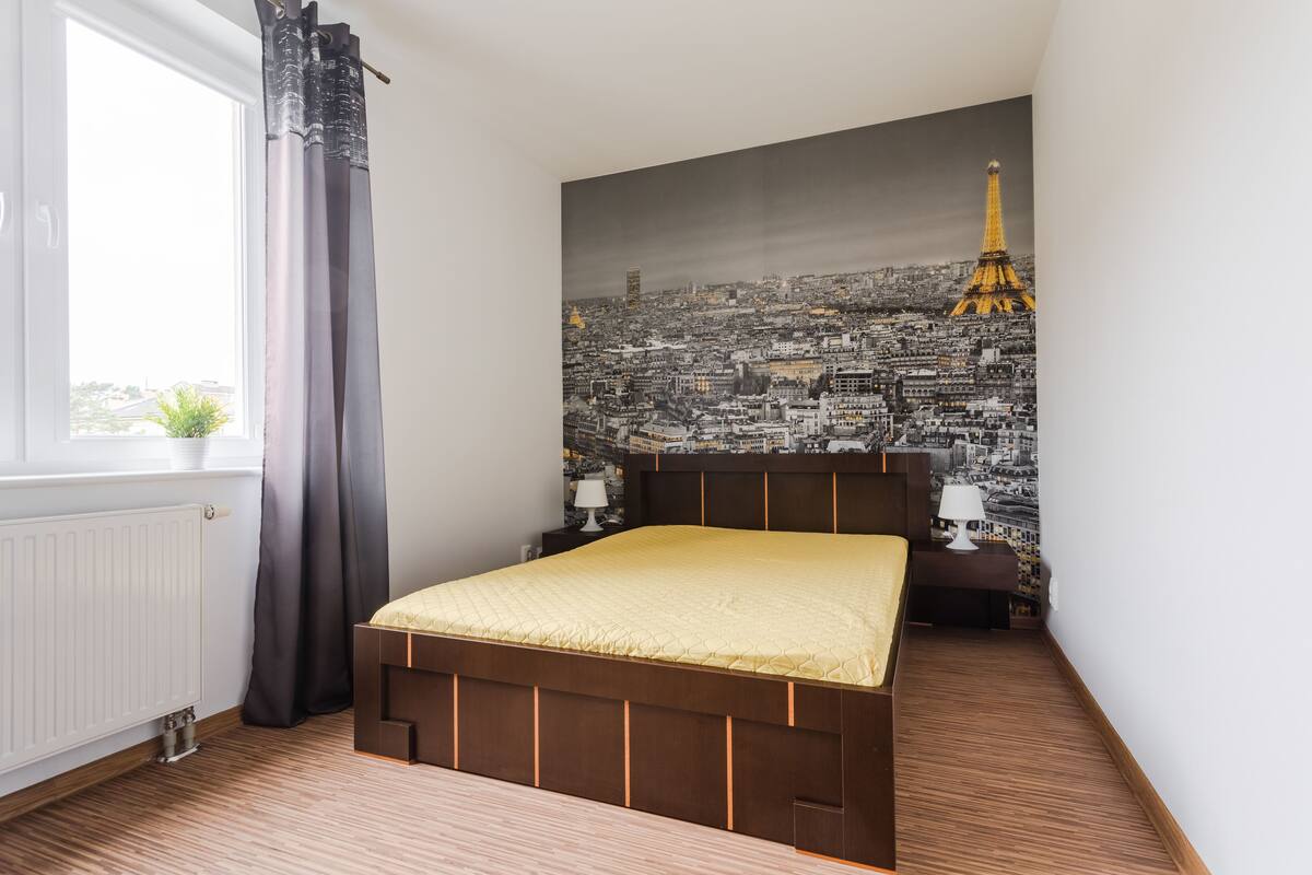 A bedroom with a bed, a window with curtains, a radiator, and a cityscape mural on the wall.