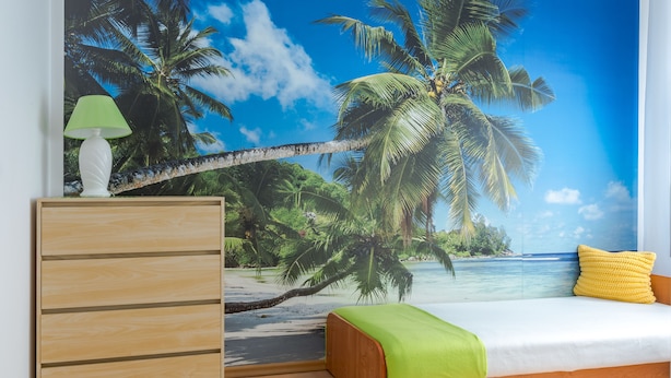 Un dormitorio con un mural de playa, un aparador de madera, una cama con almohada verde y amarilla, y una lámpara verde.