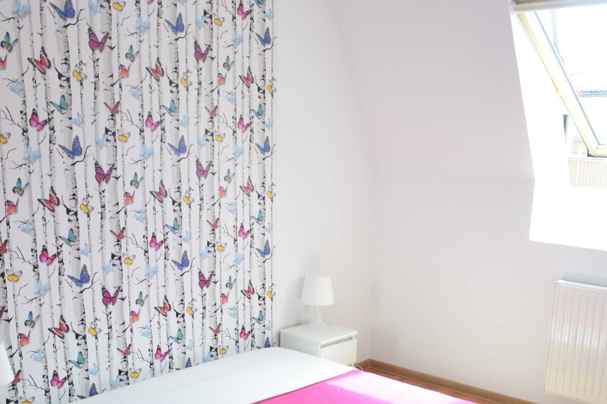 A bedroom with a bed, a nightstand, a lamp, a window, and a curtain with a butterfly pattern.
