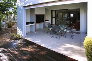 A modern outdoor patio with a glass table and chairs, a built-in grill, and a view into the house.